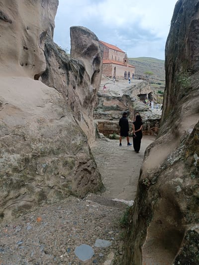 The image shows a pathway between large rock formations leading to the Uplistsikhe Cave Monastery in Georgia. In the foreground, two individuals are walking away from the camera, one in a black outfit and the other in black shorts and a black top. The pathway is dirt with scattered stones and some patches of moss and grass on the rocks. In the background, the monastery, with its red tiled roof and stone facade, is visible perched on a hillside. Several other people are scattered around the monastery and the surrounding rock structures. The sky is overcast with clouds. The environment appears to be a historical and archaeological site with natural rock formations carved into dwellings and religious structures.