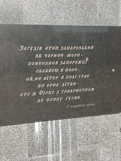 This is a close-up, eye-level photograph of an engraved stone plaque, likely a monument or memorial, set against a textured granite wall. The plaque is dark and rectangular, with white text inscribed on its surface. The text appears to be in Cyrillic script and is written in several lines, forming a poem or inscription. The bottom of the image shows a lighter-colored granite surface, possibly the base of the monument, with vertical lines suggesting it's made of multiple tiles or sections. The lighting suggests daytime, but there are no strong shadows to indicate a specific time. The overall mood is solemn and commemorative, appropriate for a public inscription. The text translates to a snippet of a Ukrainian folk song, speaking of Zaporozhian Cossacks wandering the steppe like a storm. The inscription is located in Pokrov, Ukraine.