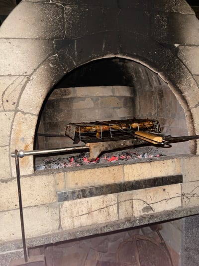 A roasting spit is centered within a brick oven in Ivankovychi, Ukraine. The spit holds a metal grill basket containing several pieces of grilled food, likely meat, that are rotating over glowing red embers. The oven's interior is constructed from light-colored bricks, many of which are stained with soot and show signs of heat damage, particularly around the arched opening. A metal rod, part of the roasting mechanism, extends horizontally through the oven. Below the oven opening, a dark, shadowed area is visible, containing what appears to be a woven basket. The exterior of the oven is also constructed from light-colored bricks, with a dark, rough stone base.