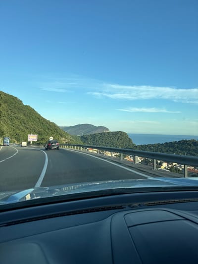 The image captures a view from a moving vehicle on a coastal highway, likely near Gornji Katun, Montenegro. The road curves to the right, with a metal guardrail separating the highway from a steep, forested hillside on the left. The hillside is densely covered in green trees.
Beyond the guardrail, the scene opens up to a panoramic view of the Adriatic Sea. Several buildings with red-tiled roofs are visible nestled among the trees on the slope overlooking the water. The sea itself is a deep blue, meeting a distant, hazy coastline dotted with more green hills. The sky is a clear, bright blue with a few wispy white clouds.
On the highway, several vehicles are present. In the foreground, the dashboard of the vehicle from which the photo is taken is visible. A black SUV is traveling ahead on the highway, with a blue truck further in the distance. A white van is also visible on the highway. A large billboard is situated on the left side of the road. The scene appears to be bathed in the warm light of either early morning or late afternoon.