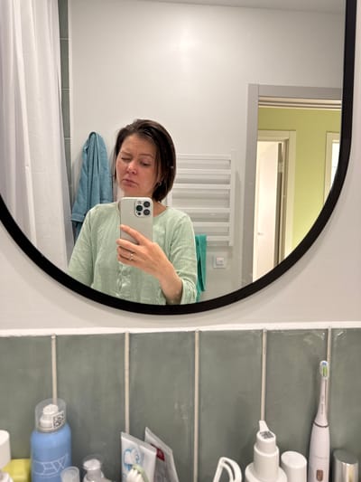 A selfie taken in a bathroom, reflected in a round mirror with a black frame. A woman wearing a light green, button-down shirt is holding a smartphone to take the picture. A white shower curtain hangs in the lower left corner. A blue towel is visible behind the woman. Behind her in the reflection, there is a white, heated towel rack. On the right side of the frame, there is a wall with large, light green tiles, holding an electric toothbrush and various toiletries. The location is presumably Люк, Russia.