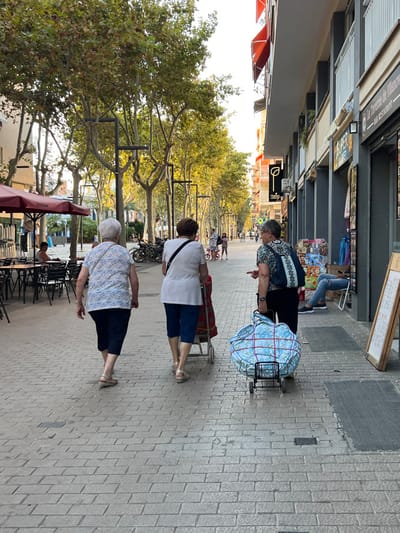 This image displays the Rambla Principal in Vilanova I La Geltrú, Spain, observed during late afternoon or early evening. The street is a pedestrian-friendly, brick-paved thoroughfare lined with mature trees, multi-story buildings, and commercial establishments.
In the foreground, three elderly women are walking away from the camera. The woman on the left wears a light-colored patterned short-sleeved top, dark capri pants, and light flat sandals. The central woman wears a white short-sleeved top, blue capri pants, and light wedge sandals, pushing a two-wheeled shopping cart with a red-patterned bag. The woman on the right, wearing a dark patterned short-sleeved top, dark pants, and dark shoes, carries a dark patterned backpack. She pushes a black two-wheeled cart carrying a large, light blue floral-patterned bag secured with red straps. This woman is observed looking to her right and gesturing with her right hand.
To the left, an outdoor seating area of a café is visible, featuring black tables and chairs under a reddish awning. Further down the Rambla, additional pedestrians, including adults and children, are present. Bicycles are parked along the left side. The right side of the street is bordered by multi-story buildings housing businesses, identified by signs such as "La Terraza de Manolo - BAR RESTAURANT" and a partial "SUPER MARKET" sign with a green leaf logo. Merchandise is displayed outside these shops. A person in blue jeans and a light shirt is seated on a white chair outside a shop on the right. Golden light filters through the tree canopy, illuminating the street.