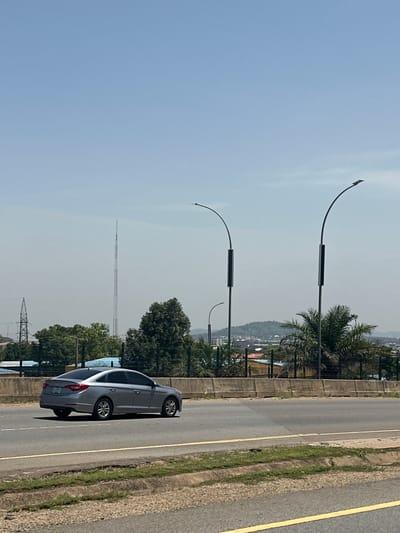 A silver sedan drives on an asphalt road in Wupa, Nigeria. The car is in the left lane, and the right lane is empty. The road is lined with a concrete barrier and vegetation. In the background, there are several tall streetlights, trees, and buildings. A tall communication tower is visible in the distance. The sky is clear and blue.
