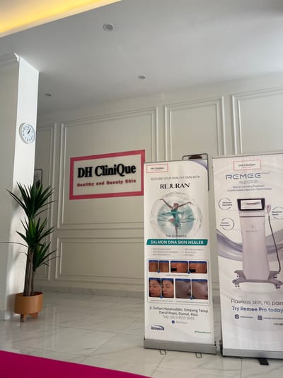 The image shows the interior of a clinic with a modern, clean aesthetic. On the wall, a sign reads "DH Clinique Healthy and Beauty Skin." To the left of the sign, a round clock is mounted on a white column. In front of the wall, there are two large standing banners. The banner on the left advertises "REJURAN" and "THE AUTHENTIC SALMON DNA SKIN HEALER," featuring images of skin treatments and before-and-after photos. This banner also displays the clinic's address as "Jl. Sultan Hasanuddin, Simpang Tetap Darul Ihsan, Dumai, Riau" and a phone number, "0823 8555 0993," along with the clinic's Instagram handle "@dhclinique." The banner on the right advertises "REMEE PRO INJECTOR" with "World-Leading Premium Comfortable Injection Technology," highlighting features like "Minimum Downtime," "Painless Treatment," and "Maximum Result." It also shows a device and contact information for the clinic, including a phone number and Instagram handle. A tall, potted plant with long green leaves stands to the left of the banners. The floor is tiled with a marble pattern, and a bright pink carpet is visible in the foreground. The walls are white and feature decorative molding. The overall environment suggests a professional and well-maintained beauty or medical spa in Dumai City, Indonesia.