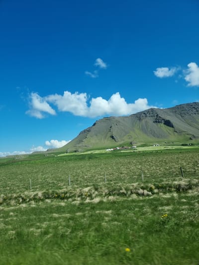 The image depicts a rural landscape under a bright blue sky with scattered white clouds. In the foreground, a vast expanse of green grass, some of it appearing slightly yellowed and windblown, stretches across the frame. Several fence posts are visible, marking a boundary within the field. Further back, the terrain rises into a green, sloping hillside covered with grass and dotted with rocks. At the base of the hill, a cluster of buildings, possibly farmhouses with red and white roofs, are situated in a green field. Several round, white objects, resembling hay bales, are also scattered in this area. The background is dominated by a large, rugged mountain with a textured, greyish-green surface, its slopes showing ridges and shadows. The overall scene is one of a serene, natural environment with evidence of human habitation. The location is Akraneskaupstaður, Iceland.