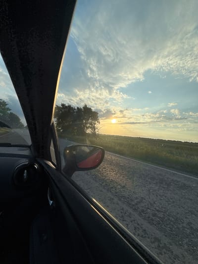 The image depicts a view from inside a car, showing a rural landscape at sunset. A road runs along the right side, bordered by a field. The sun is setting on the horizon, partially obscured by trees. A red side mirror is visible. The sky is filled with clouds. The car's interior is dark.
