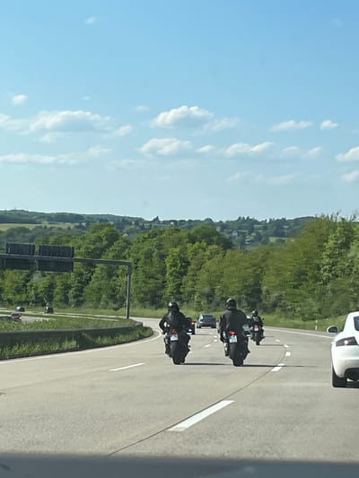 The image depicts a road in Neustadt (Wied), Germany. Two motorcyclists are riding on the road, followed by a car. The road curves through a landscape with trees and grass. The sky is blue with scattered clouds. A traffic light is visible on the side of the road. The photograph appears to have been taken from inside a vehicle.