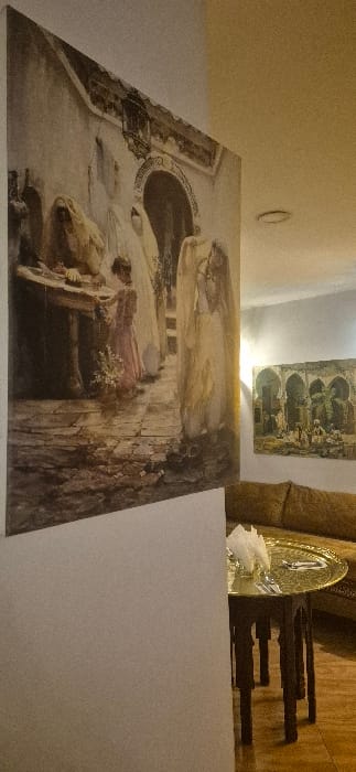 A large painting of a person pulling a cart through an arched entryway dominates the scene. The painting is hung on a white wall. Above it, a small rectangular painting depicting a building with arches is visible. To the right, a decorative plate with what appears to be napkins sits on a shelf next to some wooden objects. The room is dimly lit.