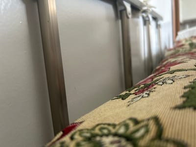 The image shows a close-up of a decorative fabric with a floral or Christmas-themed pattern, laid across what appears to be a piece of furniture with metallic legs. The fabric is primarily cream-colored with red and green floral or foliage designs. The background consists of a plain, off-white wall and several vertical metallic poles that seem to be part of the furniture or a structure behind it. The lighting is soft, suggesting an indoor setting. There are no people or animals visible in the image. The context of Kyzyl, Russia is not discernible from the visual information provided.