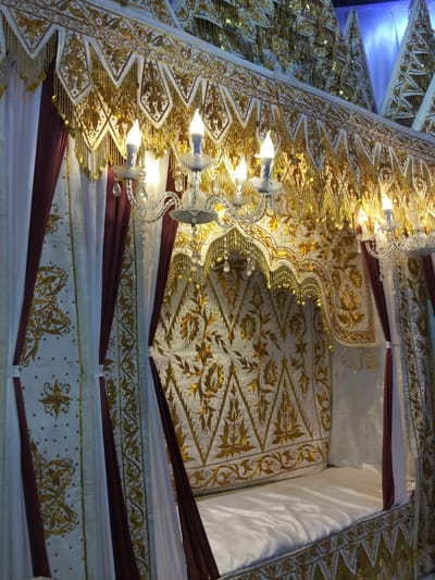 This media file shows a richly decorated ceremonial bed, likely for a wedding or important cultural event in Cottrueng, Indonesia. The bed itself is draped in white fabric, with a white mattress. The headboard and canopy are adorned with intricate gold embroidery and beading in traditional patterns. Swags of gold fabric hang from the canopy, interspersed with delicate gold chains and beadwork. Deep red curtains with gold trim are drawn partially back, revealing the opulent interior.
Two ornate chandeliers with crystal accents hang from the canopy, each with lit bulbs that cast a warm glow on the surroundings. The overall impression is one of extreme luxury and ceremonial importance. The details of the embroidery, including floral motifs and geometric borders, suggest a high level of craftsmanship. There is no visible text in the image. The lighting and the elaborate decorations suggest this is an indoor setting, possibly a decorated room or hall, prepared for a significant cultural event. The mood is celebratory and grand.