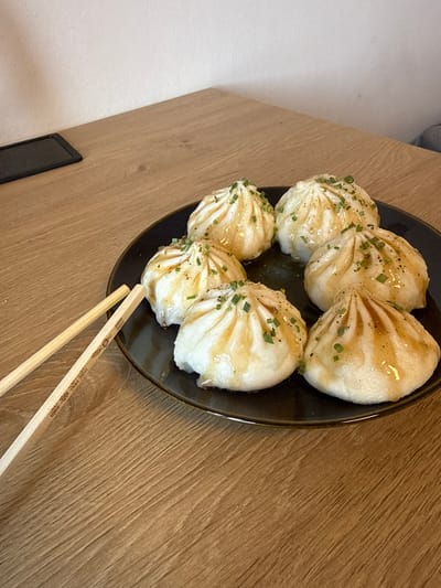 The image shows a plate of soup dumplings on a wooden table. The dumplings are round and white, with a shiny glaze and sprinkled with chopped green onions. There is a pair of chopsticks resting on the table next to the plate. The background is a white wall. The scene is indoors. The lighting suggests it is daytime. This image does not provide any information about the city of Damme, Belgium, or any other specific location.
