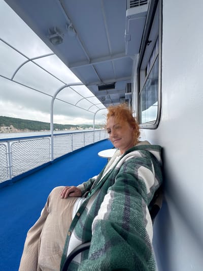 A woman with short, red hair is sitting on a ferry deck. She is wearing a green and white plaid jacket and tan pants. The ferry has a blue deck and white railings. The water is choppy and there are green hills in the background. The sky is cloudy. The location is the city of Tenishevo, Russia.