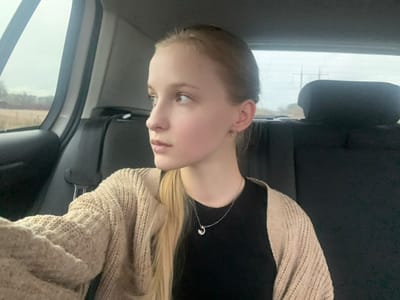The scene is situated in the city of Sonyachne, Ukraine. A young female subject, characterized by blonde hair and fair skin, is positioned in the rear passenger compartment of a vehicle. Her gaze is directed toward her left, out of the car's side window. She is attired in a black garment layered with a light beige, patterned knit cardigan. A silver-tone necklace featuring a crescent-shaped pendant is visible, along with small hoop earrings on both ears. A seatbelt is fastened across her left shoulder and chest. The vehicle's interior features dark-toned upholstery, including a headrest visible behind the subject. The exterior environment, observed through the windows, consists of an overcast sky above a flat landscape of dry, brown vegetation, with indistinct structures and power lines visible in the distance.