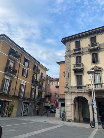 The image captures a street scene in Lecco, Italy, showing two adjacent buildings with distinct architectural styles. The building on the left side of the frame features a light yellow facade with ornate detailing around the windows and roofline. It has a large arched entranceway with a sign reading "COALVI" visible beneath the arch. A scooter is parked next to this building. The building on the right side of the frame is a darker shade of yellow and has balconies with potted plants. The sky above is blue with scattered clouds. The street is paved and appears to be in a pedestrian zone. The image is taken from a low angle, looking up at the buildings and sky.
