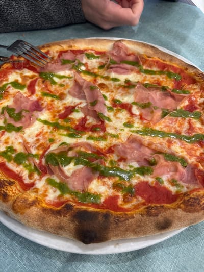 This close-up image depicts a partially consumed pizza on a white round plate, situated on a light blue textured tablecloth, within a dining establishment in Ferrara, Italy. The pizza features a well-baked, golden-brown crust with some charred spots, a red tomato sauce base, melted white cheese, and is generously topped with numerous slices of light pink cured ham (prosciutto) and a vibrant green pesto sauce drizzled in parallel lines across its surface. A silver four-tined fork is visible in the upper-left quadrant, partially embedded in a pizza slice. Above the pizza, the hands of an individual, with a gray knitted garment visible on the left arm, are positioned, indicating the ongoing act of eating.