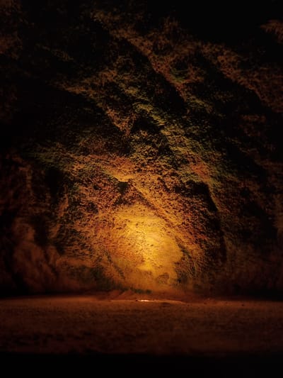 The image shows a section of a sandstone wall in a dimly lit environment. The sandstone has a rough, textured surface with variations in color, ranging from dark browns and greens in the shadows to a warm orange where illuminated by a light source. To the right, a vertical edge suggests a corner or opening, with the space beyond appearing even darker. Based on the location in Homburg, Germany, this is likely a section of the historic Schlossberghöhlen (castle mountain caves), known for their illuminated sandstone formations.