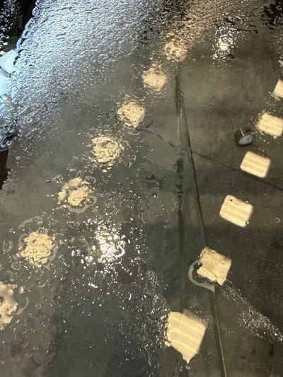 The image shows a dark, wet surface, possibly a floor, with numerous water droplets and reflections. There are several irregularly shaped, light-colored objects scattered across the surface. In the upper left, the surface appears darker and wetter. A distinct seam or line runs diagonally from the upper left to the lower right. In the right portion of the image, a series of uniformly shaped, lighter colored rectangular objects are arranged in a line. One of these objects appears to have a small, cylindrical dark object on top of it. Water is visibly running or pooling around the objects on the right. The overall scene suggests an outdoor or industrial environment where water is present, possibly in Fontevivo, Italy.