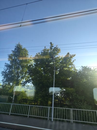 The image depicts a view from a moving vehicle, likely a train, looking out at a suburban landscape on a clear day. In the foreground, a metal railing separates the viewpoint from a paved area. Beyond the railing, lush green trees and vegetation dominate the scene. A lamppost is visible to the left of center, partially obscured by the trees. Overhead, power lines crisscross the bright blue sky. The reflections in the window suggest the presence of other vehicles or structures. The location is identified as Konobeevo, Russia.