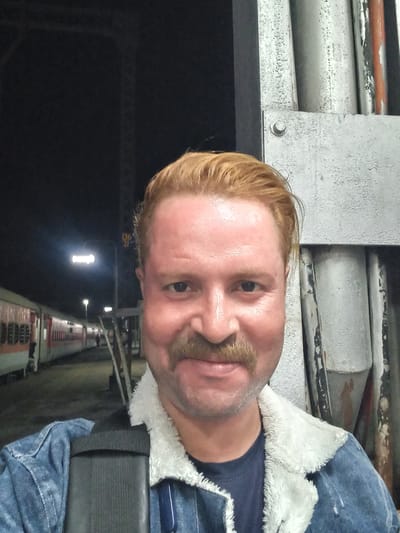 A close-up, tilted, selfie-style image shows a fair-skinned man with reddish-blond hair and a mustache. He is wearing a denim jacket with a white, fuzzy collar and a dark blue shirt. A black shoulder strap is visible. The background includes a structure with lights in the distance and a dark sky above. A grey, metallic surface is visible in the foreground. Based on the visual information available, it's impossible to determine the specific location within the city of Igatpuri, India.