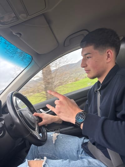 A person is inside a car, possibly the driver. The person is wearing a dark blue zip-up jacket, a seatbelt, jeans with rips at the knees, and a dark-colored smartwatch on the left wrist. The person is pointing a finger towards the windshield. The car's steering wheel is visible. The scene outside the car window shows a rural landscape with green fields and trees. The location in the city of Spahievo, Bulgaria is not discernible.