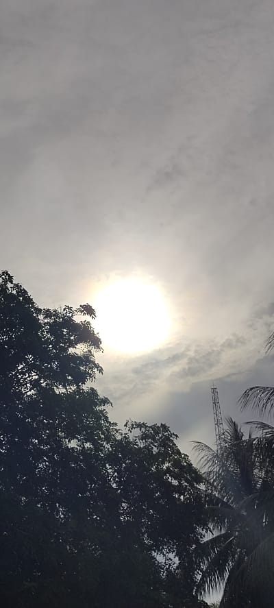 The image shows a bright sun obscured by a thin layer of clouds, creating a hazy effect. The sky is mostly overcast. The silhouette of trees is visible in the foreground. A radio tower stands in the background, right of center. The picture was taken outdoors, presumably in Aluekeujruen, Indonesia. There are no people visible in the frame.
