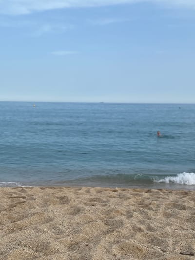 A wide shot of a beach in Vilassar de Mar, Spain, shows the foreground is a sandy beach with ripples and textures from the sand. The middle ground features the calm, blue sea with gentle waves lapping at the shore. In the distance, a person is swimming in the water, with their head visible above the surface. A yellow buoy is visible further out in the sea. The background is a clear, pale blue sky with a few wispy clouds. The overall impression is a peaceful day at the beach.