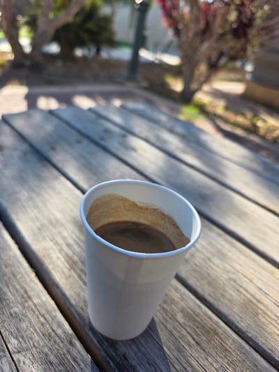 A white paper cup of coffee rests on a weathered wooden picnic table. The coffee, dark and rich, is visible at the rim of the cup. In the background, out of focus, are trees and foliage, suggesting an outdoor setting. The lighting indicates it is daytime, with shadows cast by unseen objects. The scene is located in the city of Ramat Negev Regional Council, Israel.