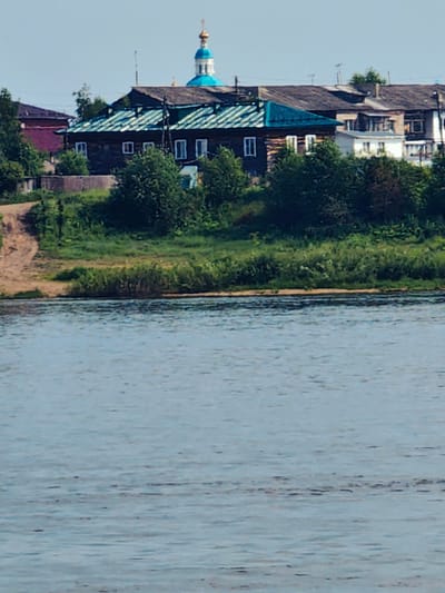 The image shows a view of the city of Yeniseysk, Russia, from across a body of water. In the foreground, the water is calm with ripples reflecting the sky. The middle ground features a lush green embankment dotted with trees and shrubs. Behind the embankment, a cluster of buildings is visible. The most prominent structure is a large wooden building with a teal-colored roof and several windows. To its right, a portion of a blue-domed church spire can be seen, topped with a golden cross. Several other buildings, including some with tiled roofs and at least one with a white facade, are also present, suggesting a residential or mixed-use area. The sky is clear and light blue. A dirt path slopes down the embankment on the left side of the image.