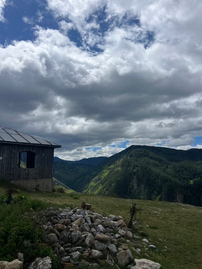 The image depicts a mountainous landscape in Giovren, Bulgaria. A small wooden structure stands in the foreground with a window facing the viewer. To the right of the structure, a pile of rocks is arranged on a grassy slope. In the background, a forested mountain rises, partially obscured by dramatic cloud formations. The sky features a mix of dark gray clouds and patches of blue.