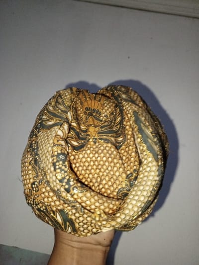 A human hand with medium-toned skin holds a folded or wrapped piece of fabric against a plain, light grey wall. The fabric, possibly a head covering or decorative textile, displays a complex pattern. This pattern includes a base of beige or light brown featuring a repetitive scale-like or small circular motif. Interwoven darker green or blue sections with gold glitter accents form stylized, curvilinear designs on the fabric. The image is set in the city of Na Sang, Laos, though no specific environmental features of the city are discernible in the background.