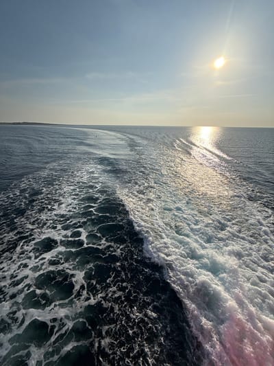 The image displays a stern view from a moving vessel in the open sea, likely in the vicinity of Kamariotissa, Greece. A prominent, frothing white wake from the vessel bisects the dark blue-green water, extending from the lower right corner towards the distant horizon. The sun, a bright circular light source, is positioned in the upper right quadrant of the sky, casting a strong, shimmering reflection across the water that stretches towards the lower right. The sky is clear, a pale blue, with a faint linear contrail visible in the upper right. On the distant left horizon, a low-lying landmass or coastline is visible. A small, dark, vertical object, consistent with a navigational buoy or marker, floats on the water in the middle ground, slightly to the left of the central wake. No individuals or other vessels are present.