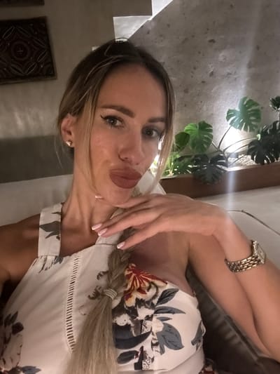The image depicts a single female adult posing for a selfie in an indoor setting within Tumbak Bayuh, Indonesia. The female subject has long, blonde hair styled in a braid extending over her left shoulder and chest. She has a light skin tone, full lips with dark reddish-brown lipstick, defined eyebrows, and dark eyeliner. A small, triangular stud earring is visible in her left ear. She wears a light-colored sleeveless top or dress with a floral pattern featuring red, grey, and brown elements, and a textured vertical stripe down the front. A gold-toned watch with a rectangular face and linked band is on her right wrist. Her left hand is raised, with fingers touching her chin, displaying manicured nails with French tips. The background features a textured, light grey wall. To the subject's left, a dark square artwork with a symmetrical pattern is mounted on the wall. To her right, a planter contains large-leafed green plants, possibly Monstera, with white decorative stones, illuminated by an unseen light source. Sections of a light-colored staircase are visible in the upper right. The overall lighting is dim and ambient.