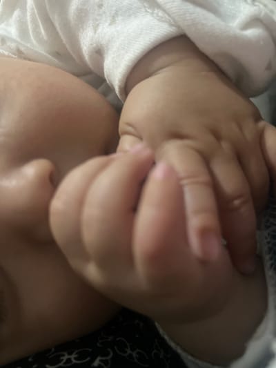 The image is a close-up, overhead shot of a baby's face and hands. The baby's left cheek and side of the head are visible on the left side of the frame, facing downwards. The baby is wearing a white garment with a subtle pattern, which is partially covering the head and body. The baby's right hand is positioned in front of the face, with fingers curled inwards and touching the cheek or mouth area. Another hand, likely the baby's left hand, is positioned over the right hand, with fingers intertwined or overlapping. The lighting is soft and appears to be artificial. The background is out of focus, but suggests a soft surface, possibly bedding. There is no visible indication of the city of Kayirma, Kyrgyzstan in the image.