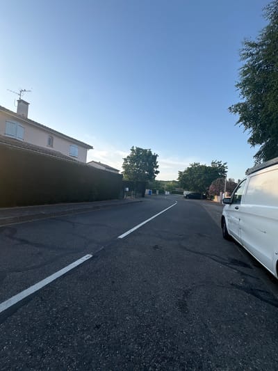 The image depicts a quiet, suburban street in Saint-Orens-de-Gameville, France, bathed in the soft light of a clear blue sky. A white van is parked on the right side of the road, its side facing the camera. The van appears to be a commercial vehicle, possibly for a tradesperson, as it has a ladder rack on its roof. The street is paved with asphalt and has a white line marking the lane. On the left side of the street, a dense, well-maintained hedge forms a barrier along the property line of a light-colored house. The house has light blue shutters on its windows and a chimney with an antenna on its roof. Further down the street, trees line the road, with some showing pinkish-purple blossoms. A black car is parked further ahead on the left side of the road. The overall scene suggests a peaceful residential area during daytime.