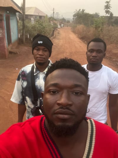 A selfie-style photograph captures three young men outdoors in Agbogugu, Nigeria. The man closest to the camera, positioned on the right, wears a red long-sleeved top with green and white stripes on the collar and shoulder. He has a short beard and an earring in his left ear. Behind him, in the center, another man is visible, wearing a plain white t-shirt. On the left side of the frame, slightly further back, a third man wears a black beanie with a white logo, possibly "Stussy," and a patterned short-sleeved shirt, along with a black cross-body bag. All three men are looking towards the camera with neutral to relaxed expressions.
The setting appears to be a rural or semi-urban environment. A reddish-brown dirt road dominates the middle ground, extending into the background. Simple buildings and lush green vegetation line the sides of the road. Distant hills or mountains are visible under a hazy sky. The natural light indicates it is daytime, likely late morning or afternoon, without strong shadows. The overall scene depicts a casual moment among friends in their local surroundings.