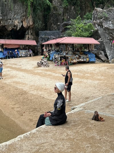 This media file depicts an outdoor scene in Tambon Krasom, Thailand. The primary subjects are several people, some of whom are interacting with market stalls. The setting appears to be a somewhat rugged area with rocky cliffs and dense vegetation, likely a natural landmark or tourist attraction.

In the foreground, two individuals are visible. One is wearing a white head covering and a black jacket, while another, wearing a black tank top and shorts, is standing and holding a phone. Behind them, a series of market stalls with pink roofs are set up, displaying various goods, including what appear to be souvenirs or local crafts. There are also tarpaulins and piles of items suggesting a bustling market environment.

The ground is sandy or gravelly, with a clear division between a lighter sandy area and a darker, more textured area, possibly indicating a path or a change in terrain. A few bags and accessories, including what looks like a Louis Vuitton bag, are placed on the ground near a rocky outcrop in the upper right portion of the image.

The overall impression is of a tourist area with commercial activity. The lighting suggests it is daytime, possibly late morning or afternoon, with no strong indications of specific weather conditions beyond it being an outdoor, possibly warm, environment. The presence of people browsing and the market stalls implies a leisurely or exploratory activity. A small sign with the number "-5" is visible on one of the stalls.