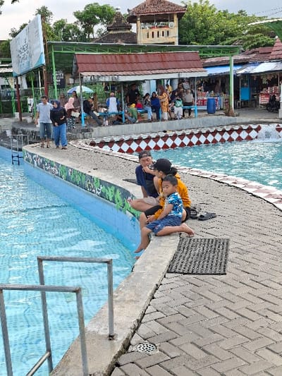 The scene is located in the town of Kecamatan Tulung, Indonesia, at latitude -7.592 and longitude 110.649. A family of three is sitting on the edge of a pool in the shade of a building. A man with a white shirt and black pants sits on a concrete ledge, next to a woman wearing a yellow top and black pants. A child wearing a blue shirt and black shorts sits in front of the man and woman. The pool is blue and the water is clear and still. The surrounding area is paved with concrete bricks and a metal handrail is positioned next to the pool. The background features several buildings and a crowd of people. The atmosphere is serene and relaxed.