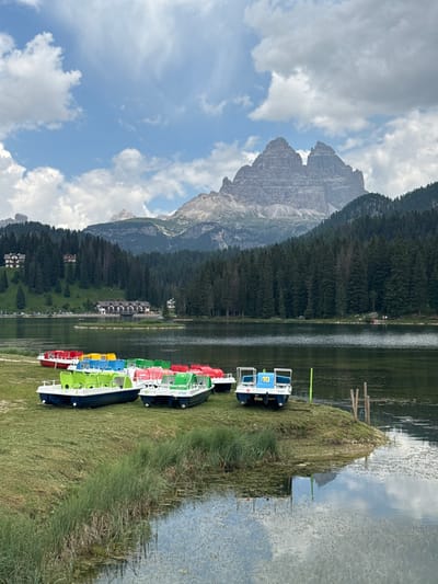The image depicts a scenic mountain lake setting in Auronzo di Cadore, Italy. The primary subjects are a row of brightly colored paddle boats lined up on a grassy shore next to a calm body of water. The boats are various shades of blue, green, yellow, and red, with some featuring numbers. A large, rugged mountain range covered in dense pine forest dominates the background, with a partly cloudy sky overhead. The scene suggests a leisurely daytime activity, likely a warm, pleasant day suitable for boating. The reflection of the sky and mountains is visible on the lake's surface. The overall impression is one of natural beauty and a tranquil vacation spot. There are no people prominently visible in the image. The only visible text appears to be numbers on the boats, such as "10" on a blue boat and "70" on a red boat.