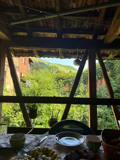 The image depicts a rustic, open-air dining area with a table set for a meal, viewed from beneath a wooden structure. In the foreground, a table is covered with a patterned tablecloth, laden with various dishes and tableware. Visible items include a patterned glass, a green bowl with a spoon, a white napkin, a small green mug, a white plate, and a bowl containing food. A dark plastic chair is partially visible behind the table.
The view extends beyond the table to a lush, green outdoor environment. The background reveals a traditional brick building with weathered walls and a terracotta-tiled roof. Surrounding the building and extending up a gentle slope is abundant greenery, including dense trees and ivy-covered walls. The landscape suggests a rural setting, with hills visible in the distance under a clear sky. The overall impression is one of a simple, outdoor meal in a village setting. This scene is likely located in the village of Shipkovo, Bulgaria, known for its natural beauty and traditional architecture.