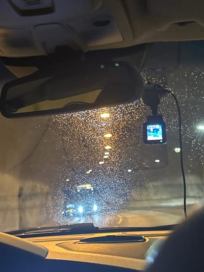 The image captures the interior of a vehicle, looking through the windshield at night. The windshield is speckled with raindrops, suggesting it is raining or has recently rained. The headlights of oncoming vehicles and streetlights create streaks of light and reflections on the wet glass, adding a dynamic, slightly blurred quality to the scene.

Prominently featured in the foreground, attached to the windshield, is a small black dashcam. Its screen is illuminated, displaying a low-resolution image, possibly of the road ahead or the interior of the car. The interior of the vehicle is dimly lit, with parts of the dashboard and a side pillar visible on the left and right, respectively. A rear-view mirror, reflecting the interior and the dashcam, is positioned on the left.

The background view through the windshield is a blurry depiction of a road at night. Lights from buildings or vehicles appear as numerous small, bright orbs scattered across the dark scene. The overall impression is one of driving in adverse weather conditions at night. The location is Røldal, Norway, which implies a potentially mountainous or rural setting common in Norway, though the specifics are obscured by the rain and darkness. No text is visible in the image.