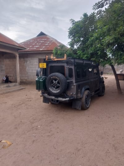 The image depicts a rural scene in Mwandege, Tanzania. A dark-colored, rugged Land Rover Defender is parked on reddish-brown dirt, angled slightly to the right, with its rear facing the viewer. The vehicle features a rear-mounted spare tire, two green jerry cans attached to the left rear, and a yellow Tanzanian license plate reading "T478 EBM" visible above the jerry cans. A roof rack is present on the vehicle's roof.
To the left of the vehicle, two individuals are visible under a covered porch area of a building constructed with light-colored cinder blocks and topped with a reddish-brown corrugated metal roof. One person is distinctly seated on the ground, leaning against the building's wall. Behind the vehicle, another structure of similar construction materials stands, characterized by a prominent high-pitched, conical reddish-brown corrugated metal roof. A large, leafy green tree is positioned to the right of the vehicle, with its foliage extending into the frame. The ground is predominantly dry, reddish-brown dirt. The sky above is overcast and grey.