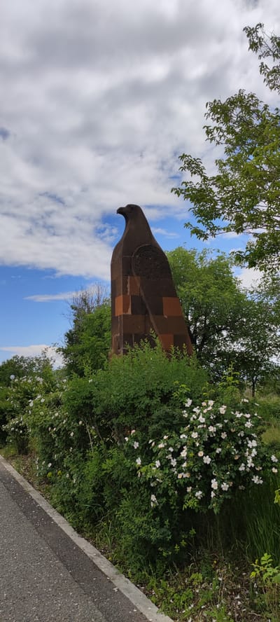 The image depicts a stone eagle statue in Araks, Armenia. The statue is brown and rectangular, adorned with an intricate circular design on its side. It stands amidst lush green vegetation and white flowering bushes along a paved road. In the background, trees and a partially cloudy sky are visible. There are no people present in the scene.