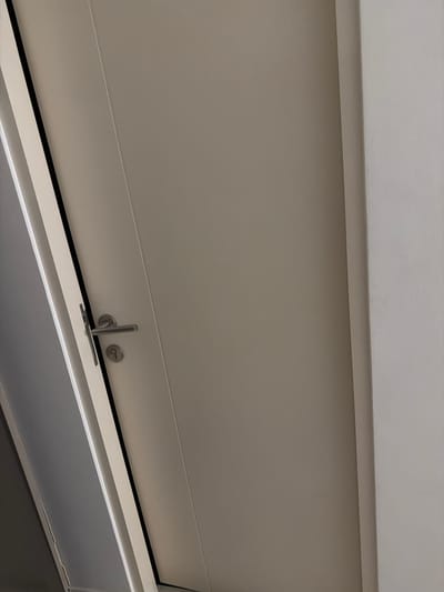 A close-up shot captures a door with a silver handle and a keyhole. The door appears to be light brown or beige, with a subtle grain pattern. To the left of the door, a portion of another door or wall is visible, with a darker, possibly greyish tone. The background on the right side of the image is a textured, off-white wall. The lighting suggests an indoor environment, possibly a hallway or a room entrance. There are no people or visible actions taking place in the scene. The image composition focuses on the details of the door and its hardware.
