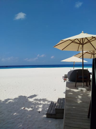 The image depicts a tranquil scene at a beach resort in Kendhoo, Maldives. The foreground features a wooden deck with two beach umbrellas, one larger and one smaller, providing shade over a dark, circular seating area. To the left of the deck, wooden steps lead down to a pristine white sand beach. The sand is marked with dappled shadows from unseen trees. The middle ground is dominated by the expansive white beach, which slopes gently towards the turquoise ocean. In the distance, a thin strip of deeper blue water marks the horizon, meeting a clear, bright blue sky. A single, small white cloud is visible in the upper left portion of the sky. On the water, far in the distance, a small dark object, possibly a boat with occupants, can be faintly seen. The overall atmosphere is one of serene tropical leisure.