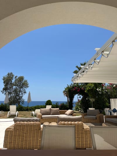 This is a photograph taken outdoors in Kissonerga Community, Cyprus, likely at a hotel or resort, showcasing a lounge area with a view of the sea. The foreground is dominated by the wicker frames of outdoor seating, including sofas and armchairs with beige cushions, arranged on a tiled patio. The middle ground features a manicured lawn with more lounge chairs and closed white umbrellas. Beyond the lawn, the blue sea stretches to the horizon under a clear blue sky. Lush greenery, including trees and flowering bushes, forms a backdrop, with an archway draped in vibrant pink bougainvillea adding a decorative element. A white retractable awning is visible on the right, extending over part of the seating area. The overall impression is one of a serene, sunny, and inviting outdoor space designed for relaxation. There are no people visible in the frame.