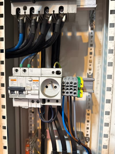 In Kallo, Belgium, this image shows a close-up view of an electrical panel. The panel contains a Merlin Gerin multi9 C60L circuit breaker, a Vigi C60 earth leakage circuit breaker with a rated residual current of 300mA and type A, and a European-style socket outlet. Several wires are connected to these components, including blue, black, brown, grey, and green/yellow wires, feeding into a grey terminal block. The electrical panel is mounted on a perforated metal backing strip, and other electrical components and cables are visible in the background, suggesting an industrial or commercial setting. The overall impression is one of electrical infrastructure within a building.