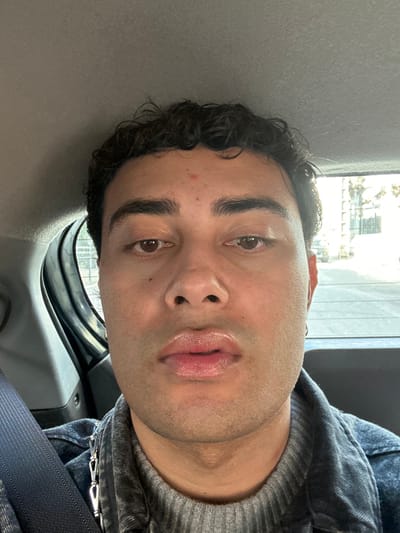 The image presents a close-up portrait of an adult individual, appearing male, seated within the rear passenger compartment of a vehicle in Neuilly-Plaisance, France. The subject has a medium skin tone, dark curly hair, and brown eyes, with a small red blemish on the forehead. The lips are full and slightly parted, exhibiting a glossy appearance. A small hoop earring is visible in the subject's right ear. The individual is dressed in a grey ribbed turtleneck sweater beneath a dark wash denim jacket. A silver-colored chain with multiple links is visible across the left side of the subject's chest, secured to the jacket. A dark blue seatbelt strap is diagonally positioned across the left shoulder and chest. The subject maintains a direct gaze towards the camera with a neutral expression. The vehicle's interior is visible, featuring a grey headliner and grey plastic trim of the rear left door. Through the vehicle's windows, an outdoor daylight scene is discernible, depicting a street with blurred buildings, vehicles, and trees, indicative of an urban or suburban setting.