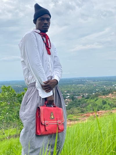 A medium shot captures a Black man standing on a grassy hill overlooking a sprawling, green cityscape in Amaigbo, Nigeria, under a cloudy sky. He is wearing a pointed black beanie, a loose white button-down shirt with its sleeves rolled up, and flowing grey pants. Around his neck, he sports a red scarf with black beaded accents. In his hands, he holds a bright red satchel with gold trim and a crest. The background reveals a panoramic view of Amaigbo, showcasing numerous buildings nestled amongst lush vegetation and rolling hills, with a visible construction site featuring orange earth and heavy machinery in the mid-ground. The foreground is dominated by tall, vibrant green grass swaying in the breeze.