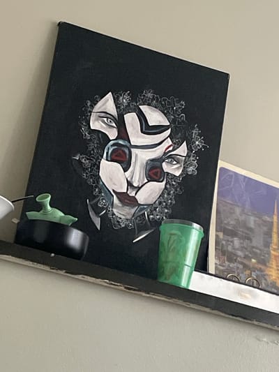 A black canvas features a stylized painting of a face with white, angular features outlined in black and red. The eyes are open, with blue irises. The face is partially surrounded by delicate white flowers. To the right of the canvas, a bright green plastic cup is resting. Further to the right and below the cup is a black object with a light green nozzle, possibly a spray bottle cap. Behind the canvas, a portion of a larger item with a metallic sheen and two rings is visible, along with a purple and white patterned surface. The overall scene appears to be indoors, possibly on a shelf or wall. There is no visible text in the image.