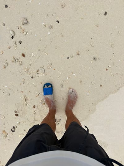 A person stands on a sandy beach, presumably on Fulidhoo Island, Maldives, given the context. The view is from slightly above, looking down at the person's legs, bare feet, and lower body. One foot is bare, covered in sand, while the other wears a blue sandal, also partly covered in sand. The person wears dark-colored shorts. The sand is light-colored and grainy, with small fragments of coral and shells scattered across the surface. The edge of the water is visible at the top edge of the image.